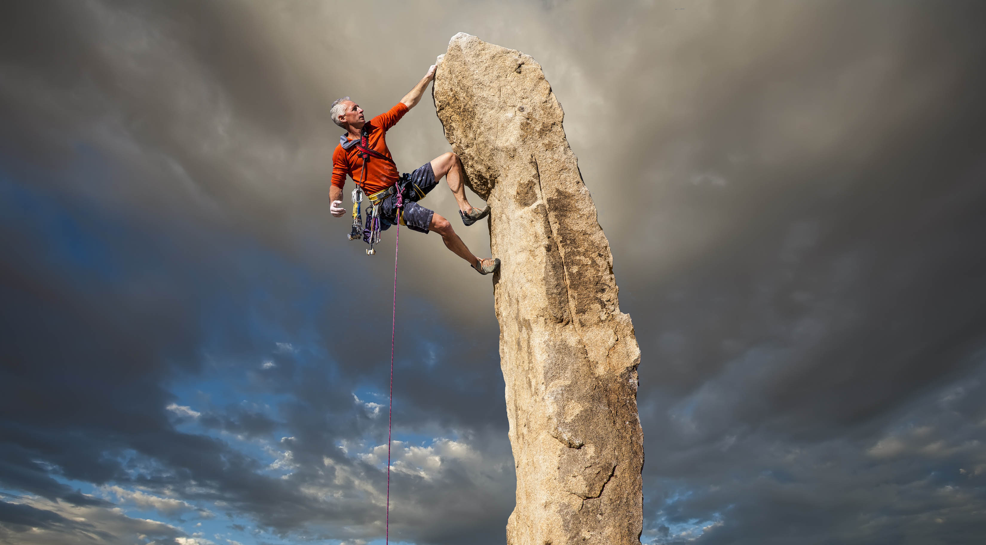 Older fit man climbing the top of a thin mountain, sky in the background is ominous Older fit man climbing the top of a thin mountain, sky in the background is ominous